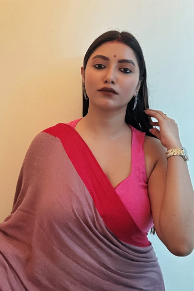 Anjali Housewife in a traditional pink and red saree stands in front of a light-colored wall. She has long straight hair,