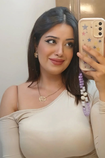 A woman stands in front of a mirror taking a selfie with her smartphone. She is dressed in a long beige off-shoulder dress that fits closely to her body.
