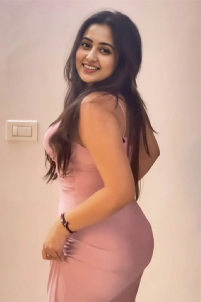 A woman in a light pink dress standing indoors, smiling and posing with her hand in her hair.